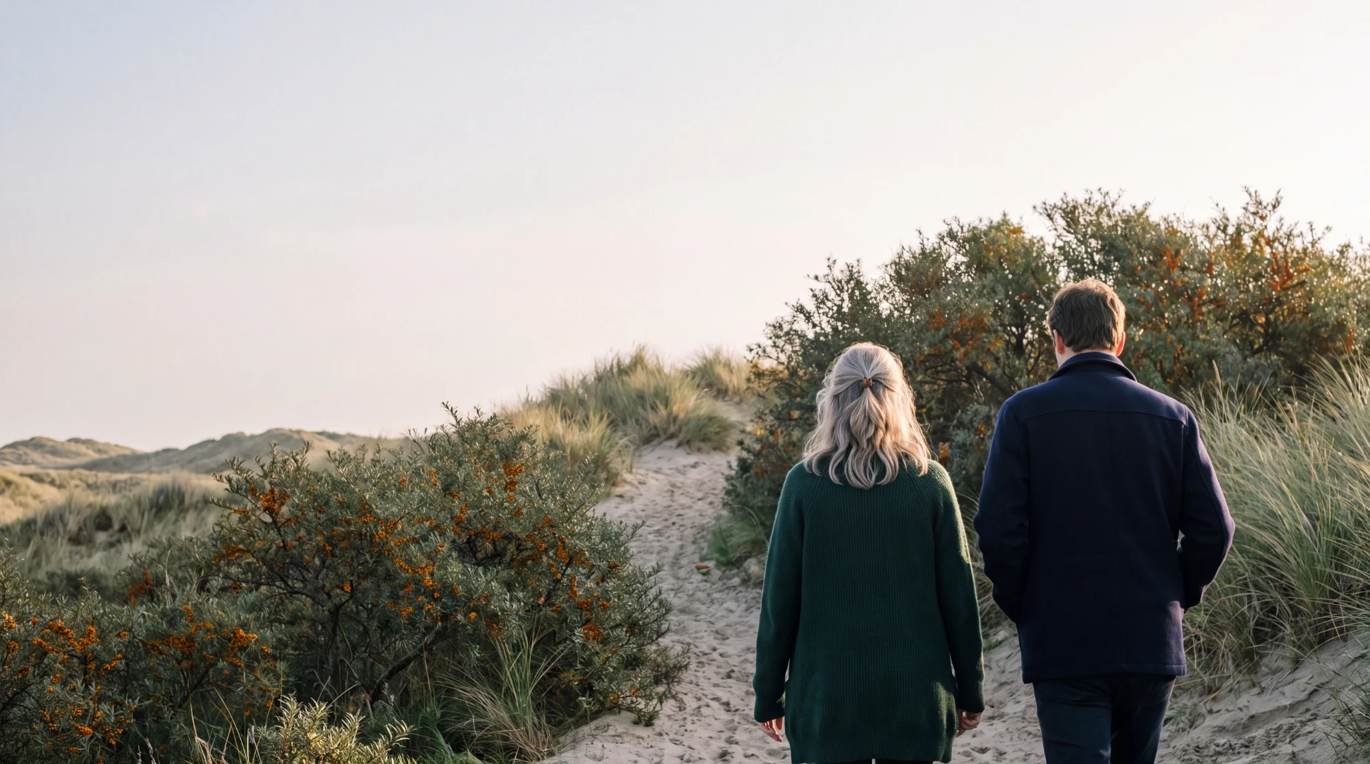 Ann en een cliënt wandelen naast elkaar over een zandpad door de duinen tussen De Haan en Wenduine, van achteren gezien, tussen duindoorn met oranje bessen.